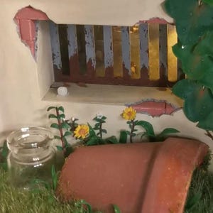 May include: A miniature garden scene with a broken wall revealing a window with gold and blue bars. A terracotta pot, glass jar, and yellow flowers are in the foreground. Green grass and foliage add to the garden theme.