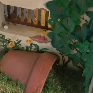 May include: A miniature garden scene featuring a terracotta pot, yellow sunflowers, and green foliage. The pot is partially buried in the grass. A decorative wall with a gold and brown painted design is in the background.