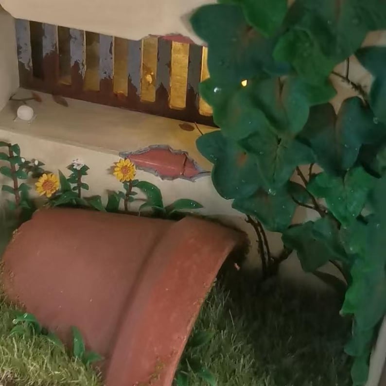 May include: A miniature garden scene featuring a terracotta pot, yellow sunflowers, and green foliage. The pot is partially buried in the grass. A decorative wall with a gold and brown painted design is in the background.