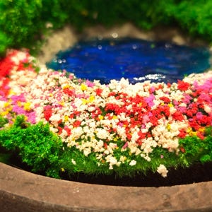 May include: A miniature garden scene featuring a blue pond surrounded by colorful flowers and green foliage. The flowers are a mix of red, white, pink, and yellow. The scene is set in a wooden bowl, creating a tranquil miniature landscape.