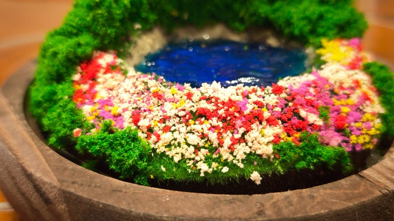 May include: A miniature garden scene featuring a blue pond surrounded by colorful flowers and green foliage. The flowers are a mix of red, white, pink, and yellow. The scene is set in a wooden bowl, creating a tranquil miniature landscape.