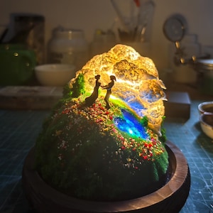 May include: A miniature illuminated sculpture of a couple holding hands on a grassy hill with colourful flowers. The sculpture has a glowing, textured rock formation and a blue and green stream. It is set in a wooden base.