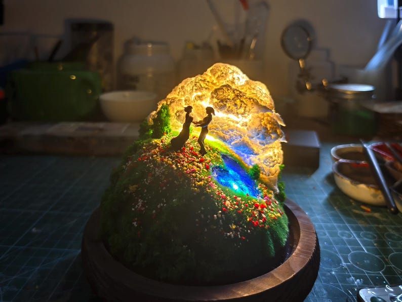 May include: A miniature illuminated sculpture of a couple holding hands on a grassy hill with colourful flowers. The sculpture has a glowing, textured rock formation and a blue and green stream. It is set in a wooden base.