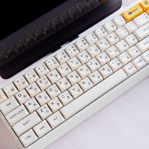 May include: A white and yellow mechanical keyboard with Japanese characters and English letters. The keyboard has a compact layout with a white frame and yellow accent keys. The keyboard is angled towards the viewer, with a black laptop in the background.