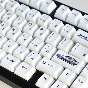 Peut inclure: Clavier mécanique blanc avec lettrage noir et caractères japonais. Le clavier comporte des touches personnalisées avec un dessin de voiture et du texte japonais. Un cadran argenté est sur le coin supérieur droit.