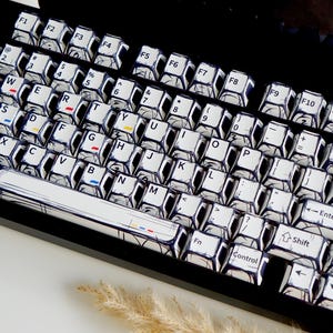 May include: A black mechanical keyboard with white keycaps featuring a unique comic book style design. The keys are labeled with black text and accented with red, yellow, and blue details. A circular dial is on the right side.