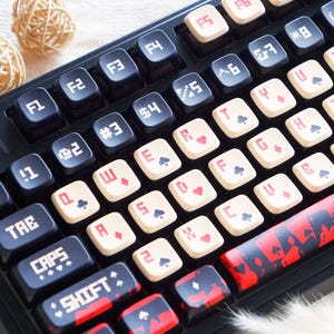 May include: A black mechanical keyboard with custom keycaps. The keycaps feature playing card symbols, with red and black designs. The keyboard also has a red and black space bar, and a keycap with a cartoon image.