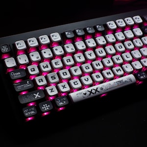 Pode incluir: Teclado mecânico preto com teclas brancas e retroiluminação rosa. As teclas são marcadas com letras e símbolos brancos. O teclado tem um design personalizado com teclas exclusivas e um apoio para os pulsos com texto e gráficos.