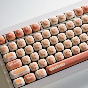 May include: A white mechanical keyboard with custom keycaps. The keycaps are a light peach color with cartoon faces and letters. The function keys are a darker peach color. The space bar and other keys have food-themed illustrations. The keyboard is angled.