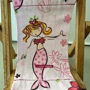 Puede incluir: Una silla de playa rosa con un dise&ntilde;o de sirena. La sirena lleva un sujetador de concha rosa y una cola de lunares rosa y blanca. La silla es de madera y tiene una tela a rayas rosa y blanca.
