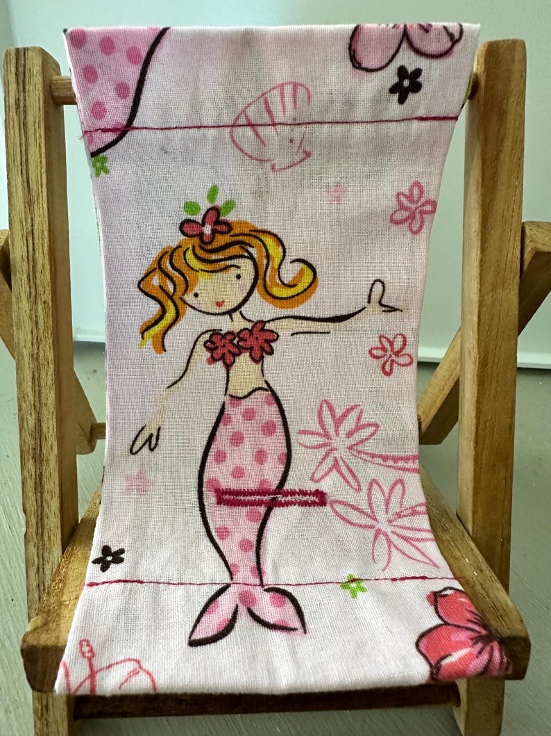 Puede incluir: Una silla de playa rosa con un dise&ntilde;o de sirena. La sirena lleva un sujetador de concha rosa y una cola de lunares rosa y blanca. La silla es de madera y tiene una tela a rayas rosa y blanca.