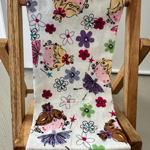 Puede incluir: Una peque&ntilde;a silla plegable de madera con un asiento de tela blanca que presenta un patr&oacute;n caprichoso de hadas y flores en tonos de rosa, morado, verde y rojo.