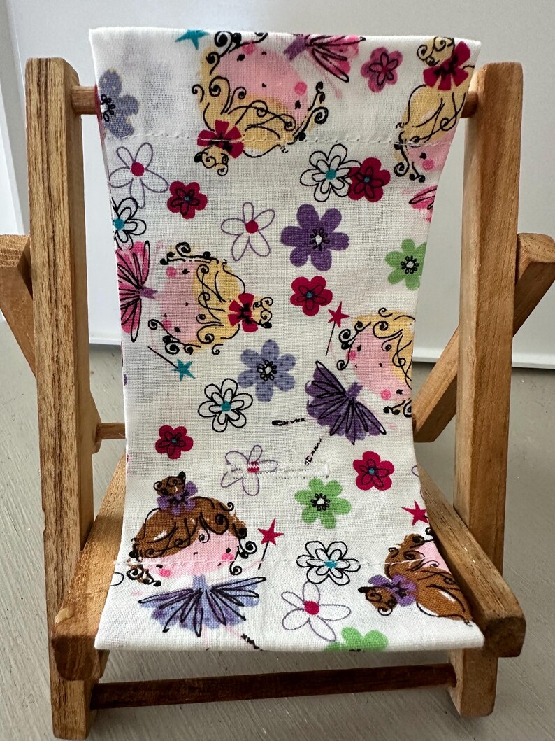 Puede incluir: Una peque&ntilde;a silla plegable de madera con un asiento de tela blanca que presenta un patr&oacute;n caprichoso de hadas y flores en tonos de rosa, morado, verde y rojo.
