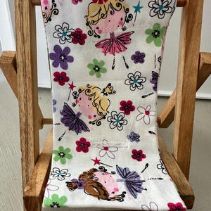 Puede incluir: Una silla plegable de madera con un asiento de tela blanca con un patr&oacute;n repetitivo de flores coloridas y hadas de dibujos animados.