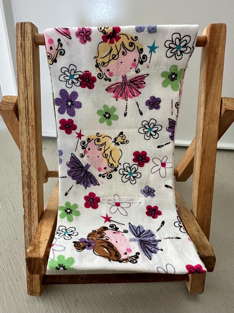 Puede incluir: Una silla plegable de madera con un asiento de tela blanca con un patr&oacute;n repetitivo de flores coloridas y hadas de dibujos animados.