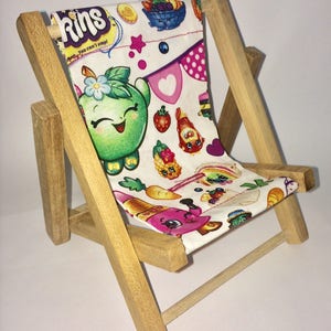 Puede incluir: Una peque&ntilde;a silla plegable de madera con un asiento de tela blanca con un patr&oacute;n colorido de personajes de dibujos animados y el texto "Shopkins You can't stop!"