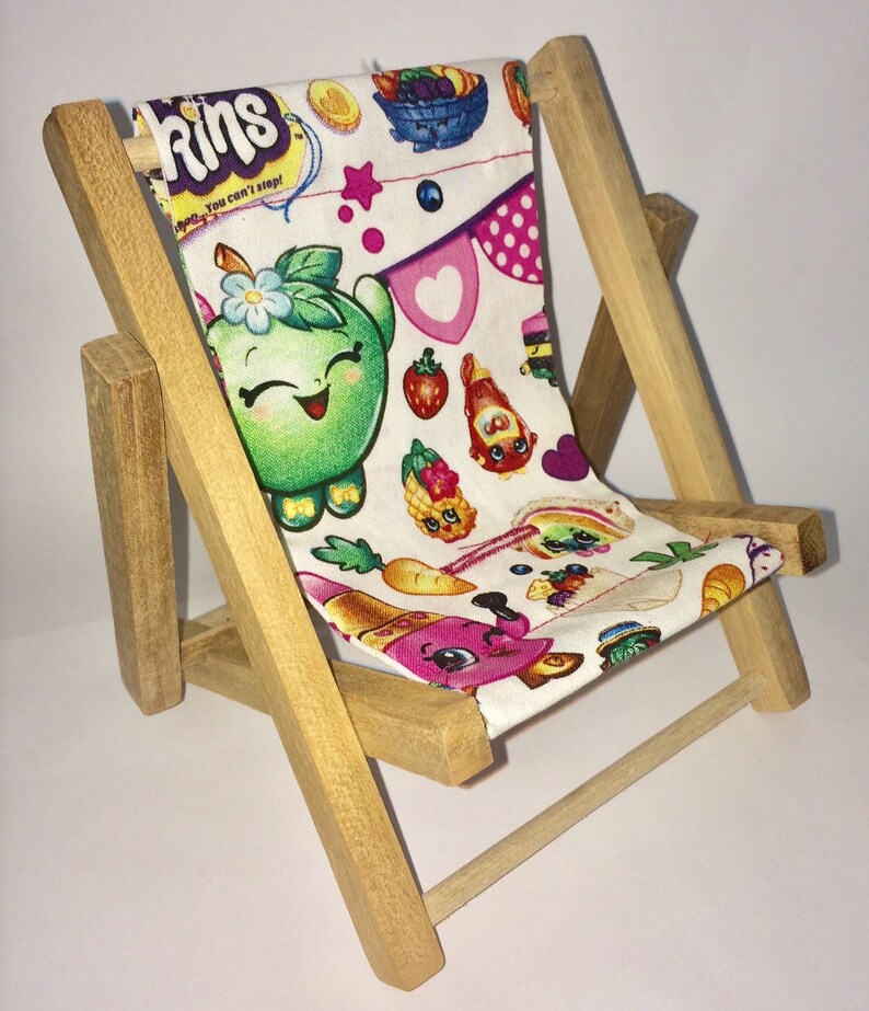 Puede incluir: Una peque&ntilde;a silla plegable de madera con un asiento de tela blanca con un patr&oacute;n colorido de personajes de dibujos animados y el texto "Shopkins You can't stop!"