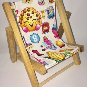 Puede incluir: Una silla plegable de madera con un asiento de tela blanca con un patr&oacute;n colorido de personajes de dibujos animados y alimentos. Los personajes son de la marca Shopkins e incluyen una galleta, una mazorca de ma&iacute;z, una lata de refresco y una escoba.