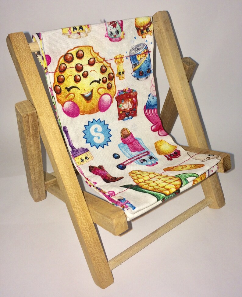 Puede incluir: Una silla plegable de madera con un asiento de tela blanca con un patr&oacute;n colorido de personajes de dibujos animados y alimentos. Los personajes son de la marca Shopkins e incluyen una galleta, una mazorca de ma&iacute;z, una lata de refresco y una escoba.