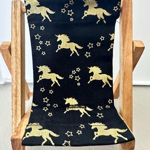 Puede incluir: Una peque&ntilde;a silla plegable de madera con un asiento de tela negra con un patr&oacute;n de unicornio dorado y estrellas.