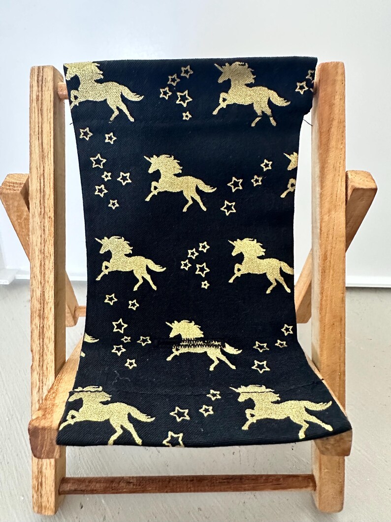Puede incluir: Una peque&ntilde;a silla plegable de madera con un asiento de tela negra con un patr&oacute;n de unicornio dorado y estrellas.