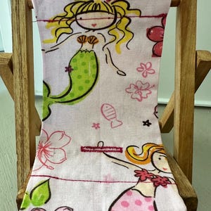 Puede incluir: Una silla de playa de tela rosa y verde con un dise&ntilde;o de sirena. La silla est&aacute; hecha de madera y tiene un asiento y un respaldo de tela. La tela presenta sirenas, flores y peces.
