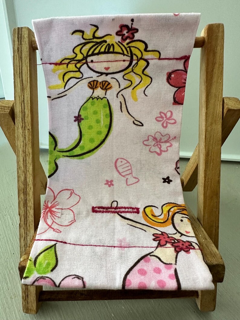 Puede incluir: Una silla de playa de tela rosa y verde con un dise&ntilde;o de sirena. La silla est&aacute; hecha de madera y tiene un asiento y un respaldo de tela. La tela presenta sirenas, flores y peces.