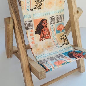 Puede incluir: Una silla de playa de madera en miniatura con un asiento de tela que presenta un patr&oacute;n colorido de Moana y Pua de la pel&iacute;cula de Disney Moana. La tela es blanca con acentos azules, naranjas y marrones.