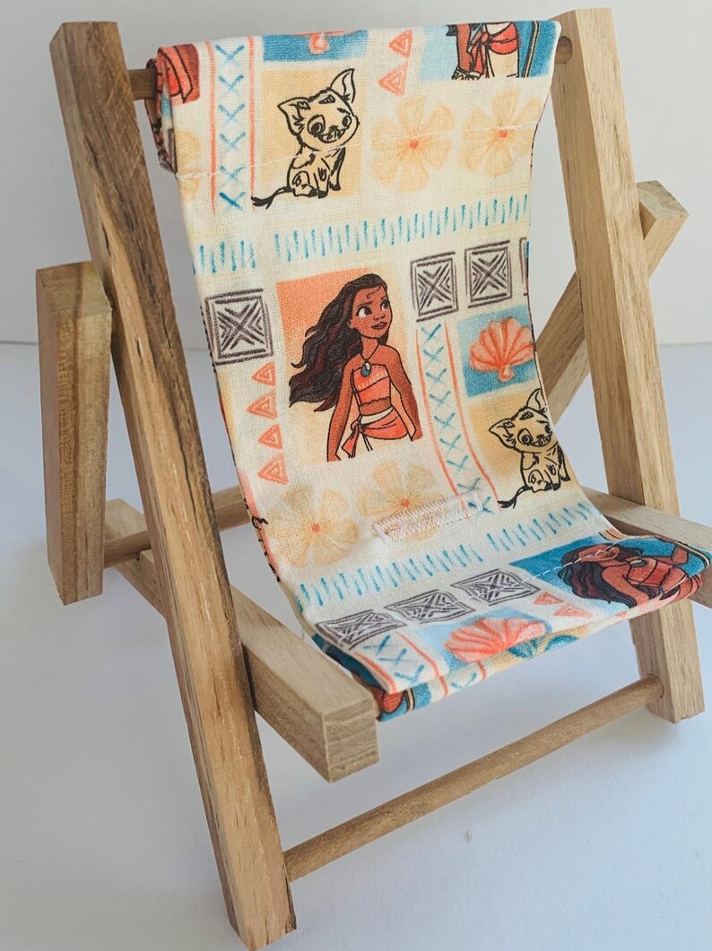Puede incluir: Una silla de playa de madera en miniatura con un asiento de tela que presenta un patr&oacute;n colorido de Moana y Pua de la pel&iacute;cula de Disney Moana. La tela es blanca con acentos azules, naranjas y marrones.