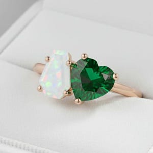 May include: A gold ring with two gemstones. One is a white, pear-shaped opal, and the other is a heart-shaped, emerald-green stone. The ring is set in a white jewelry box.