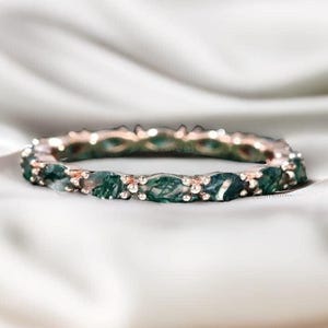 Puede incluir: Un anillo de oro rosa con una banda de piedras de ágata musgosa verde de forma ovalada. El anillo se encuentra sobre un fondo blanco y sedoso, resaltando el contraste entre el metal y las piedras preciosas. Una joya.