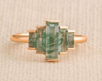 May include: A gold ring featuring five rectangular, moss-green gemstones. The gemstones are set in a horizontal arrangement, with a polished gold band. The ring's design is modern and elegant, suitable for everyday wear or special occasions.