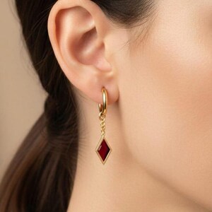 May include: Gold hoop earrings with a dangling red diamond-shaped charm. The earrings are worn on a person's ear, showcasing the jewelry's design and color. The red charm is set in a gold frame.