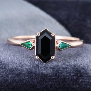 Puede incluir: Anillo de oro rosa con una gran piedra central negra hexagonal flanqueada por dos piedras verdes triangulares. El anillo tiene una banda delgada y un acabado pulido, mostrando un diseño moderno.