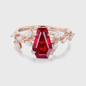 May include: Rose gold ring featuring a large, faceted, red gemstone in a coffin shape. The ring is adorned with small, white, marquise-cut stones and leaf-like details on the band, creating an elegant and unique design.