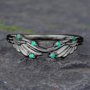 May include: A black ring with a wing design, featuring small, round, emerald-green gemstones. The ring's wings curve upwards, creating an open design. The ring is set against a dark, textured surface.