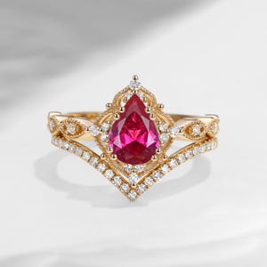 Peut inclure: Ensemble de bagues en or avec une pierre rouge en forme de poire au centre, entourée de petits diamants blancs. La bague présente un design orné et une bande assortie en dessous, créant un effet superposé.