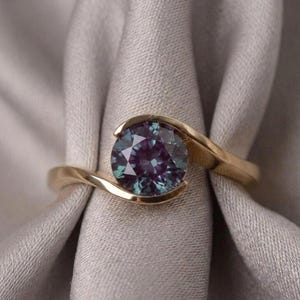 May include: A gold ring featuring a round, faceted, color-changing gemstone that displays hues of deep purple, teal, and blue. The band has a smooth, polished finish and curves around the central stone in a bypass-style setting.