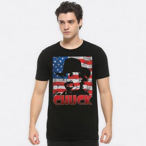 May include: Black t-shirt featuring a graphic of a cowboy with an American flag background and the name "CHUCK" in red. The design is distressed, giving it a vintage look. The shirt is a classic crew neck.