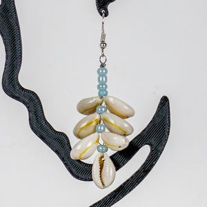 May include: A dangling earring featuring a silver-toned hook, light blue spherical beads, and multiple white cowrie shells arranged in a vertical cluster. The earring is displayed on a black, abstract, curved, 3D-printed plastic stand.