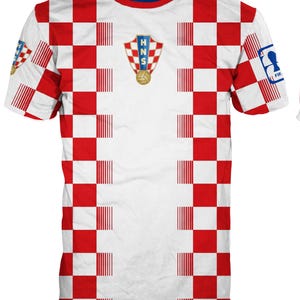 May include: A short-sleeved t-shirt featuring a red and white checkered pattern across the front and sleeves. The center chest displays the HNS crest with a soccer ball. A blue FIFA logo patch is visible on the right sleeve and a small crest on the left.