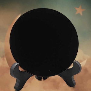 May include: A large, circular, black object is presented on a black wooden stand. The stand has a decorative, curved design. The background features a celestial theme with a star and a crescent moon.