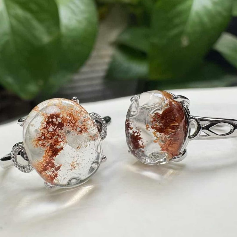 May include: Two silver rings featuring clear, oval gemstones with reddish-brown inclusions. The rings have intricate detailing around the settings and on the bands. The gemstones' patterns are unique. The rings are a delicate design.