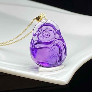 May include: A purple amethyst pendant necklace featuring a smiling Buddha figure. The pendant is carved from a translucent, polished gemstone and hangs from a delicate gold chain. The necklace is displayed on a white surface.