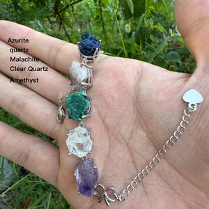 May include: A silver bracelet featuring azurite, quartz, malachite, clear quartz, and amethyst gemstones. The bracelet has a chain with a heart-shaped clasp and a bow-shaped charm. The gemstones are in silver settings.