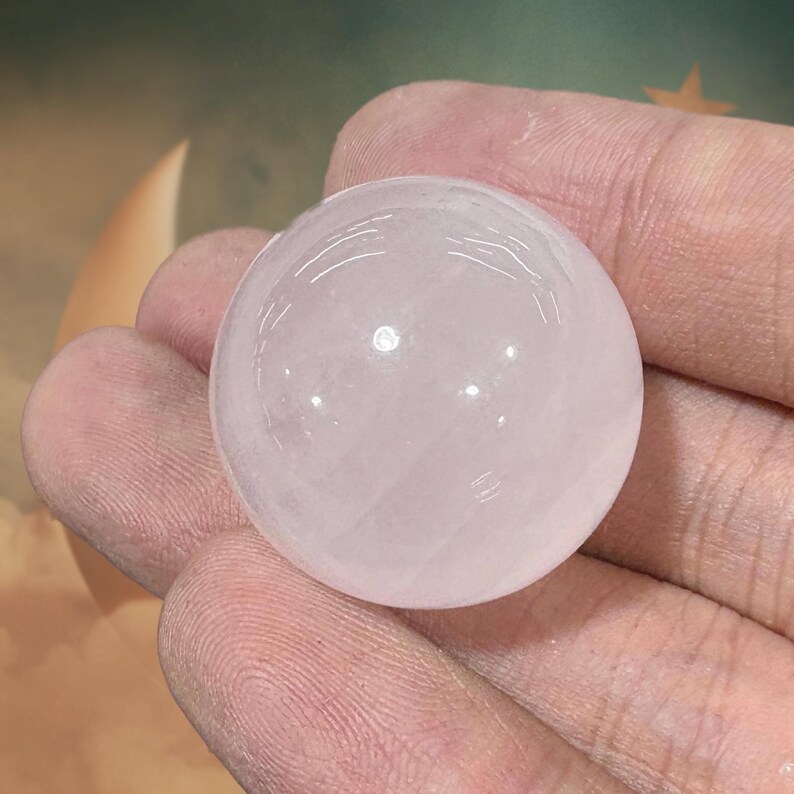 May include: A translucent, spherical rose quartz crystal ball. The polished sphere is held in a hand, showcasing its smooth surface and soft pink hue. The crystal reflects light, creating subtle highlights.