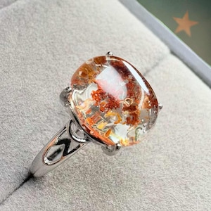 Könnte beinhalten: Silberring mit einem großen, ovalen Edelstein. Der klare Stein hat rote, orange und gelbe Einschlüsse. Der Ring hat ein dekoratives Band mit offenem Design. Ein einzigartiges Schmuckstück mit einem auffälligen, farbenfrohen Edelstein.