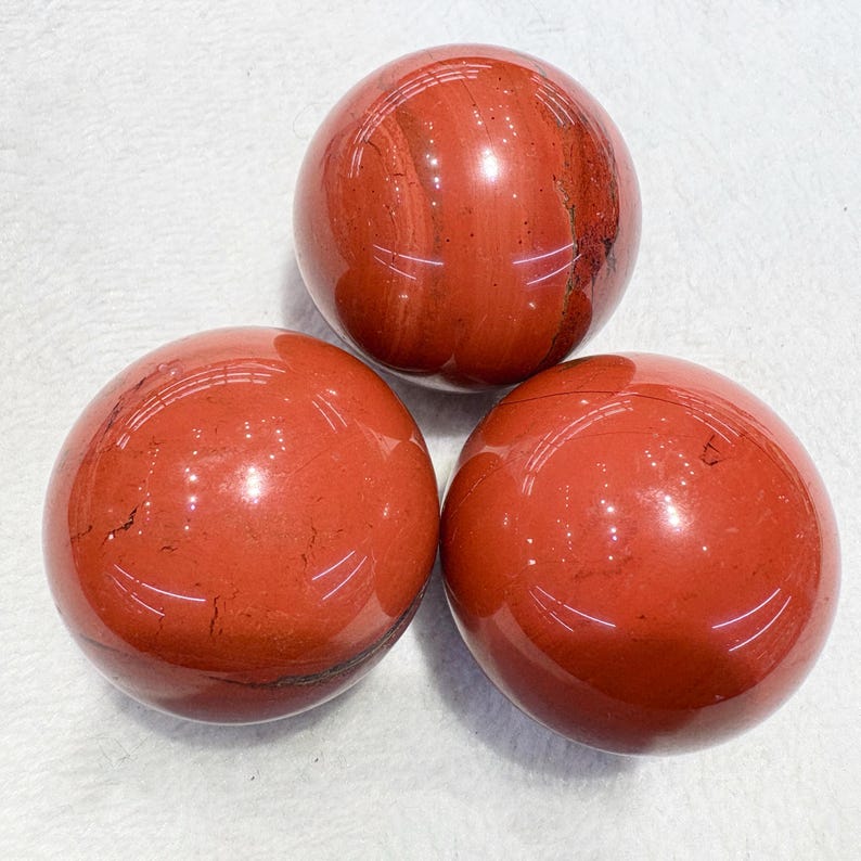 May include: Three polished red jasper spheres. The spheres have a smooth, glossy surface with natural veining and variations in color. The spheres are arranged in a triangular pattern against a white background.