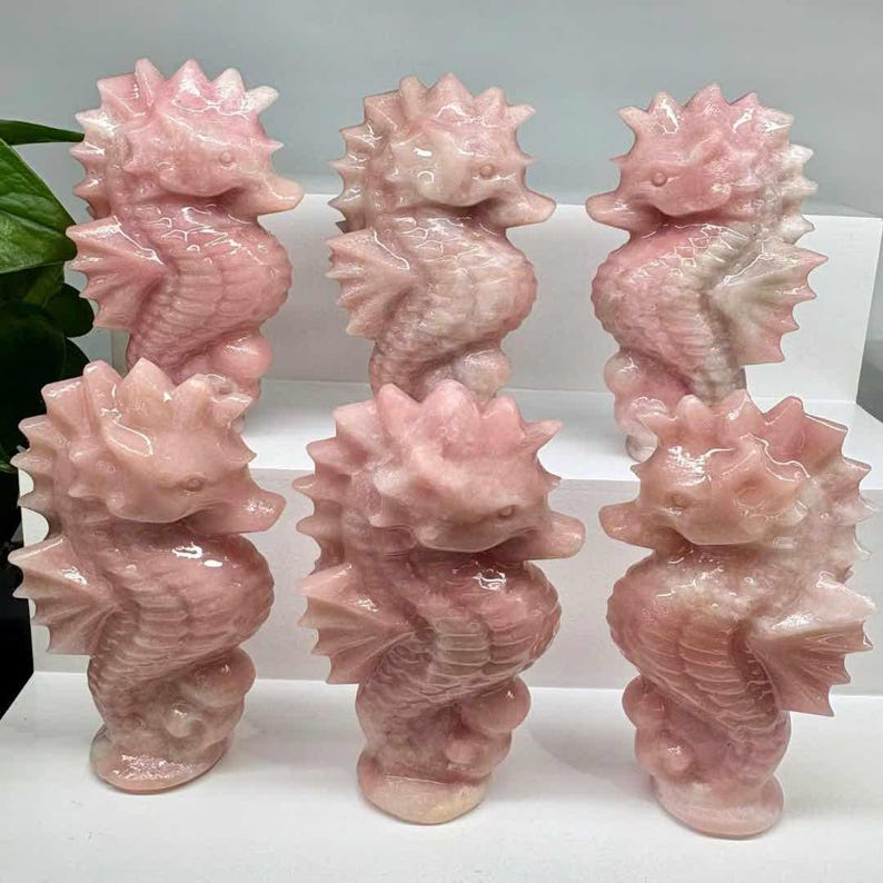 May include: Six pink crystal seahorse figurines. Each seahorse is carved with detailed fins, a spiky dorsal fin, and a curved body. The figurines are displayed on a white surface.