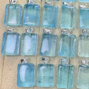 May include: A collection of rectangular, light blue pendants with silver-colored bails. The pendants have a translucent, glassy appearance, and the color varies slightly between each piece. The pendants are arranged closely together, suggesting they are for sale.